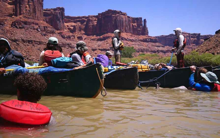 A group of canoes are tethered together while floating in calm water. There are a couple of people in the water. In the background, there are tall, red, canyon walls. 
